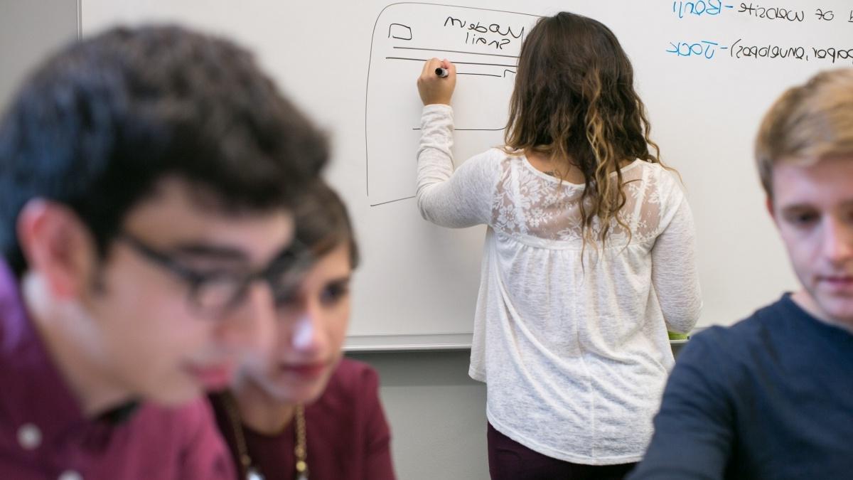 One student sketches on a whiteboard while others sit together in discussion