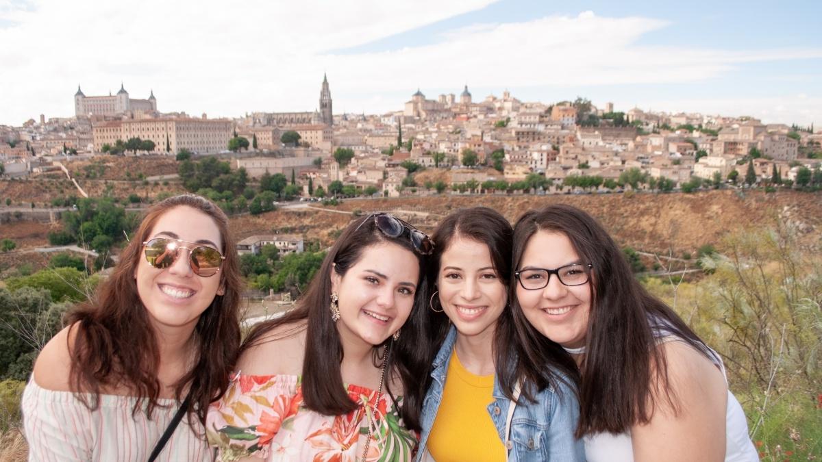 四个年轻女子微笑着，远处可以看到一座古老的西班牙小镇 